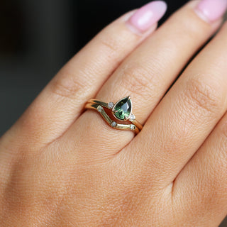 Three stone yellow gold ring with pear cut green sapphire and white round diamond accents stacked with curved wedding band with three round diamonds, on hand