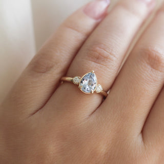 Three stone engagement ring with a light blue pear cut sapphire in yellow gold, on hand