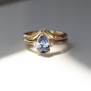 Three stone yellow gold ring with pear cut blue sapphire and white round diamond accents stacked with pointed wedding band with three white round diamonds, front view