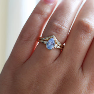 Three stone yellow gold ring with pear cut blue sapphire and white round diamond accents stacked with pointed wedding band with three white round diamonds, on hand