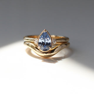Three stone yellow gold ring with pear cut blue sapphire and white round diamond accents stacked with large wave style curved wedding band, front view