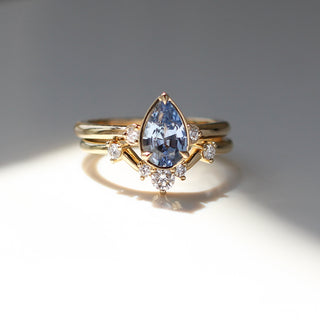 Three stone yellow gold ring with pear cut blue sapphire and white round diamond accents stacked with curved wedding band with five round white diamonds, front view