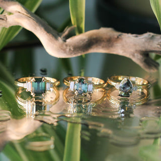Trilogy Oval Green Sapphire Ring with Emerald and Pear Cut Sapphire Rings on a mirror with reflections of branches and flowers detailed front view