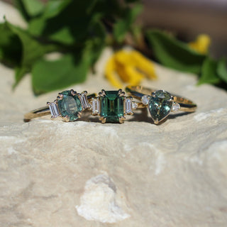 Trilogy Oval Green Sapphire Ring stacked with Emerald and Pear cut rings in sunlight on a stone front view
