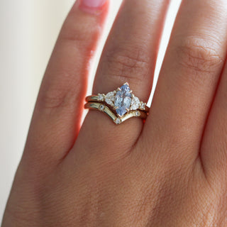 Yellow gold ring with a light blue marquise cut sapphire and white diamond clusters stacked with a pointed wedding band with three round white diamonds, on hand