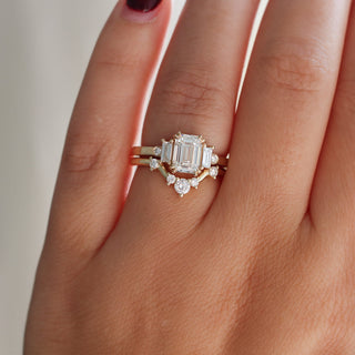 Emerald cut lab diamond ring with baguette and round diamond side stones in yellow gold stacked with curved diamond wedding band, on hand