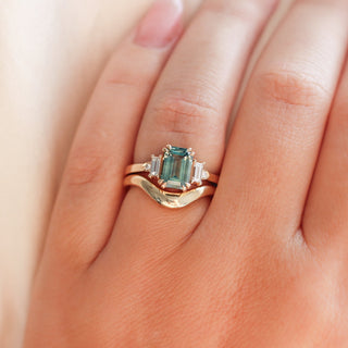 Emerald cut teal sapphire ring with baguette and round diamond side stones in yellow gold stacked with curved wedding band, on hand