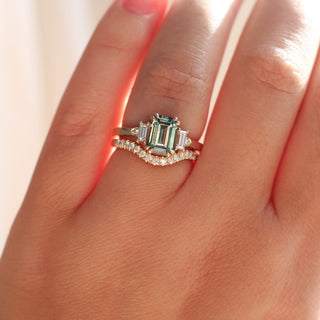 Emerald cut teal sapphire ring with baguette and round diamond side stones in yellow gold stacked with curved diamond pave wedding band, on hand