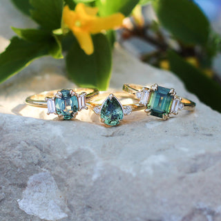 Misceo Emerald Cut Green Sapphire Ring stacked with oval and pear cut sapphire rings on a stone in bright sunlight front view