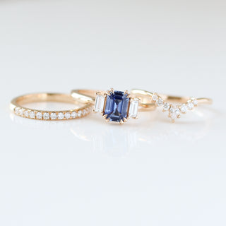 Blue emerald cut sapphire and diamond ring on white background laying beside two diamond wedding bands, front view