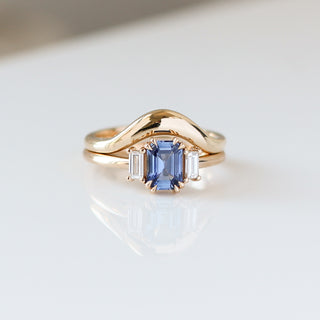 Blue emerald cut sapphire and diamond ring on white background stacked with large curved gold wedding band, front view