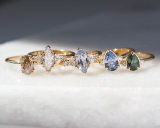 Five gold rings with marquise and pear-cut gemstones in various colors, arranged on a white textured surface with a dark gray background.