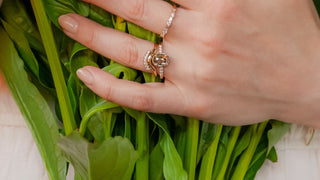 Two hands wearing gold rings with diamonds and gemstone designs, resting on a bouquet of flowers.