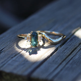 Yellow gold ring with an elongated emerald cut teal sapphire in a 5-stone design with white diamonds beside a wave style yellow gold wedding band, front view on wood