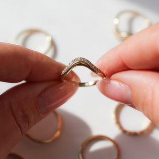 Hand holding a curved gold wedding band with diamond accents against a light background