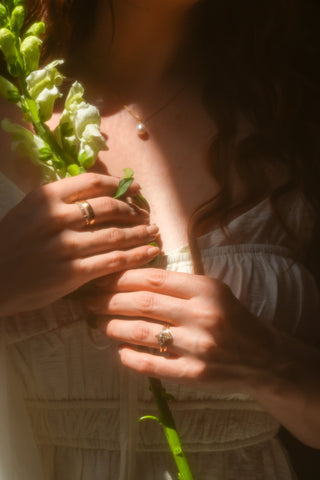 Person holding a flower with sunlight filtering through, wearing a pearl necklace and ring.