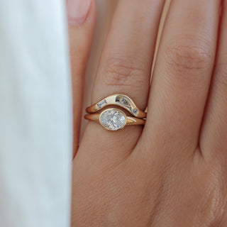 Yellow gold ring with a bezel set white oval cut diamond with a curved yellow gold wedding band with assorted white diamonds, on hand