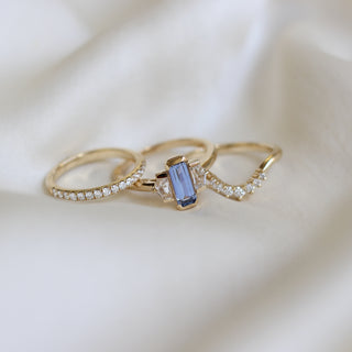 Three stone yellow gold ring, with elongated emerald cut blue sapphire half bezel set and shield shaped white sapphire side stones in half bezel settings in between two yellow gold white diamond wedding bands, front view