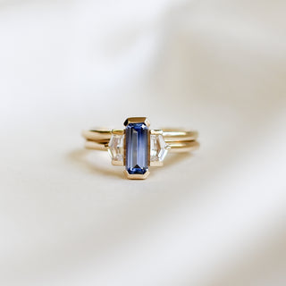 Three stone yellow gold ring, with elongated emerald cut blue sapphire half bezel set and shield shaped white sapphire side stones in half bezel settings stacked with a plain yellow gold straight wedding band, front view