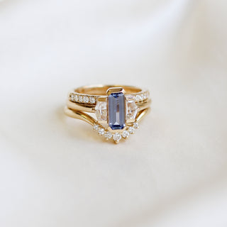 Three stone yellow gold ring, with elongated emerald cut blue sapphire half bezel set and shield shaped white sapphire side stones in half bezel settings stacked with two yellow gold white diamond wedding bands, front view