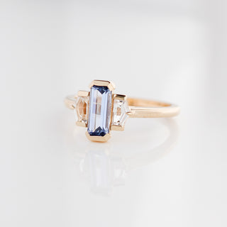 Three stone yellow gold ring, with elongated emerald cut blue sapphire half bezel set and shield shaped white sapphire side stones in half bezel settings, quarter view