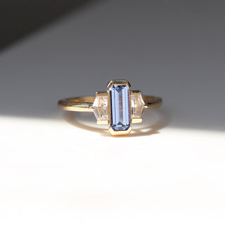 Three stone yellow gold ring, with elongated emerald cut blue sapphire half bezel set and shield shaped white sapphire side stones in half bezel settings, quarter view