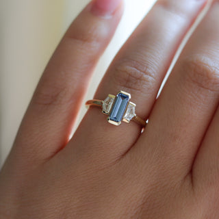 Three stone yellow gold ring, with elongated emerald cut blue sapphire half bezel set and shield shaped white sapphire side stones in half bezel settings on hand, top view