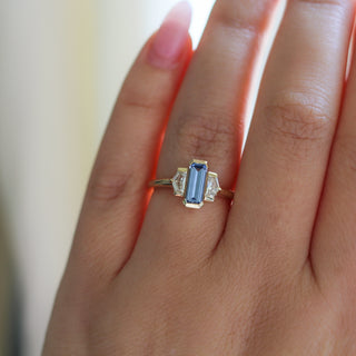 Three stone yellow gold ring, with elongated emerald cut blue sapphire half bezel set and shield shaped white sapphire side stones in half bezel settings on hand, top view