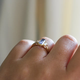 Three stone yellow gold ring, with elongated emerald cut blue sapphire half bezel set and shield shaped white sapphire side stones in half bezel settings on hand, side view