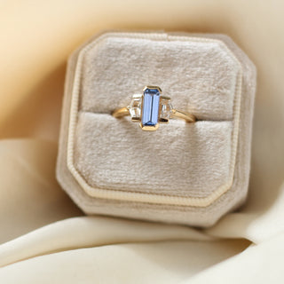 Three stone yellow gold ring, with elongated emerald cut blue sapphire half bezel set and shield shaped white sapphire side stones in half bezel settings in ring box, top view