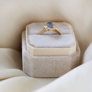 Three stone yellow gold ring, with elongated emerald cut blue sapphire half bezel set and shield shaped white sapphire side stones in half bezel settings in ring box, side view
