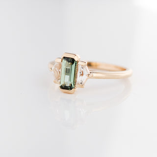 Three stone yellow gold ring, with elongated emerald cut green sapphire half bezel set and shield shaped white sapphire side stones in half bezel settings, quarter view
