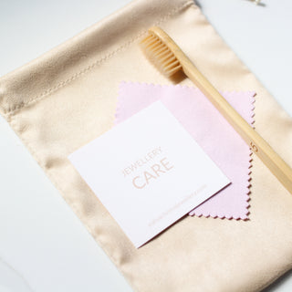 A jewellery cleaning kit with a pink polishing cloth, a recyclable bamboo brush, and a card reading 'JEWELLERY CARE' on top of a beige velvet pouch