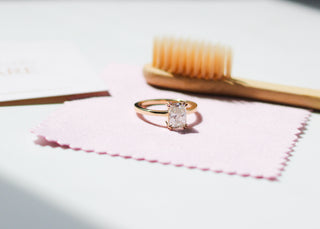 A jewellery cleaning kit with a pink polishing cloth, a recyclable bamboo brush, and a card reading 'JEWELLERY CARE' with a rectangular white diamond solitaire ring on top of the cleaning cloth