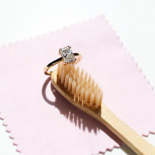 A jewellery cleaning kit with a pink polishing cloth and a recyclable bamboo brush, with a rectangular white diamond ring perched on the end of the bamboo brush