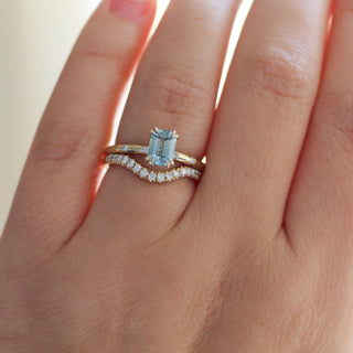 Yellow gold ring with a light blue emerald cut sapphire with yellow gold curved wedding band with white diamonds, on hand