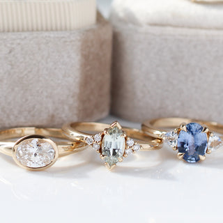 Three yellow gold rings side by side, one with a bezel set white oval cut diamond, one with a light green marquise sapphire, and one with a blue oval cut sapphire, with cream ring boxes in the background 