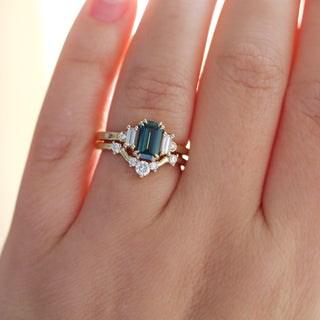 Yellow gold ring with a teal emerald cut sapphire and four white diamond accents and a yellow gold curved wedding band with round white diamonds, on hand