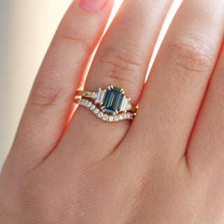 Yellow gold ring with a teal emerald cut sapphire and four white diamond accents and a yellow gold curved wedding band with round white diamonds, on hand