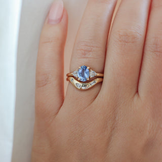 Yellow gold ring with a blue oval cut sapphire and two white trillion cut diamonds with a yellow gold curved wedding band with white diamonds, on hand