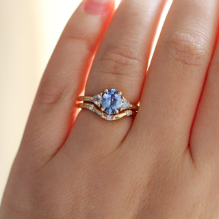 Yellow gold ring with a blue oval cut sapphire and two white trillion cut diamonds with a yellow gold curved wedding band with round white diamonds, on hand