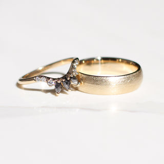 Two yellow gold wedding bands, one wide band with a beach texture and one thin curved crown band with salt and pepper marquise diamonds and round white diamonds. 