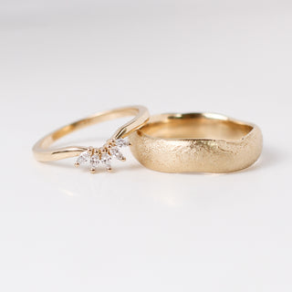 Two yellow gold wedding bands, one wide band with an organic beach texture and one thin curved band with a crown of marquise diamonds. 