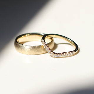 Two yellow gold wedding bands, one wide band with a beach texture and one thin curved band with pavee diamonds.  