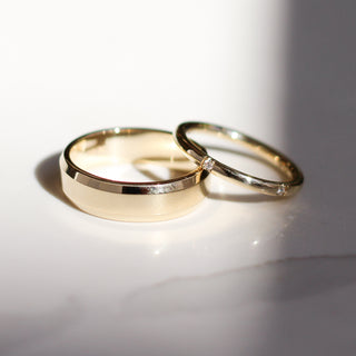 Two yellow gold wedding bands, a wide band with detailing on one edge and a thin band with two diamonds. 